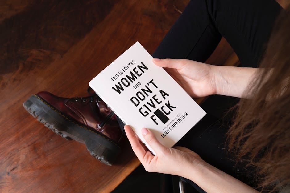 A person seated on a wooden floor holding a white paperback book titled 'This is for the Women Who Don't Give a F***' by Janne Robinson. The individual is wearing dark pants and brown leather boots, with the shoe visible in the foreground. The person's hands grasp the book, with the thumb on one side and fingers on the other, showing the title clearly. The setting suggests an indoor environment, consistent with a home or apartment, and the image is related to home relocation or packing processes, as indicated by the context of house removals and moving services provided by Man with Van Foots Cray.