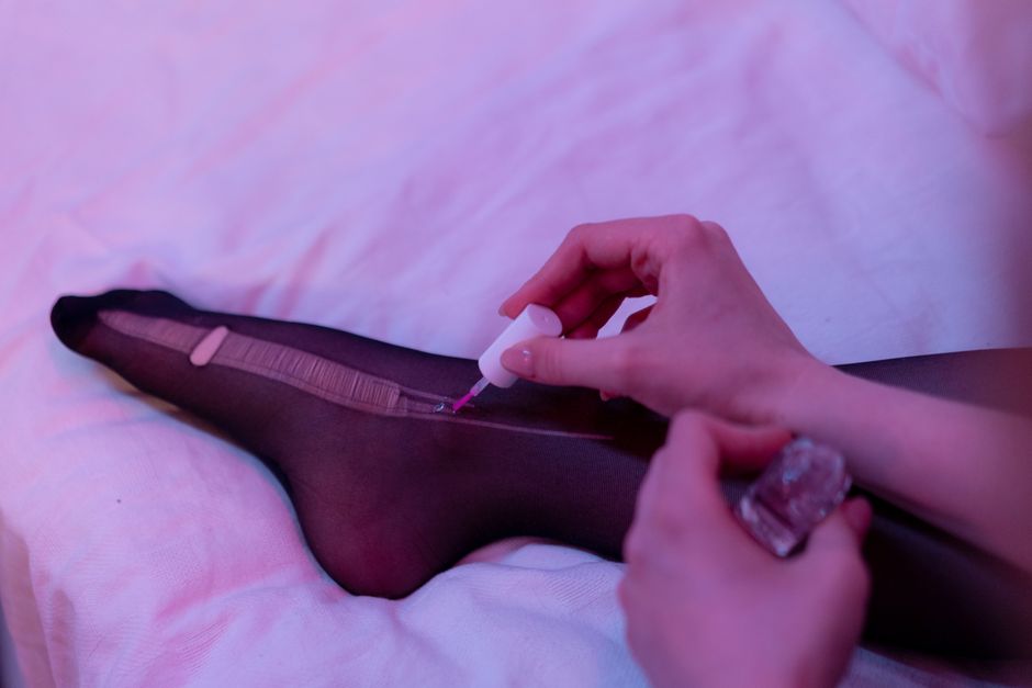 A person's hand is seen applying clear nail polish to a toenail on a foot lying on a light-colored, soft fabric surface. The foot is covered in sheer black tights, with the toes and part of the foot v
