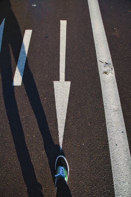 A close-up view of a black and white sneaker worn by a person standing on a paved street surface during a home relocation process. The person is positioned along the white directional arrow painted on the asphalt, indicating a parking or loading zone near a residential property. To the left, there are additional parking lines and shadows cast by nearby objects, possibly vehicles or street furniture. The pavement surface is dark with some small debris and minor cracks. This scene captures part of the loading process for furniture transport, with the arrow guiding the movement of items and vehicles involved in the house removals service provided by Man with Van Foots Cray. The area appears well-lit with natural daylight, and the scene is part of a typical moving day where parking and loading logistics are crucial for efficient home relocation.