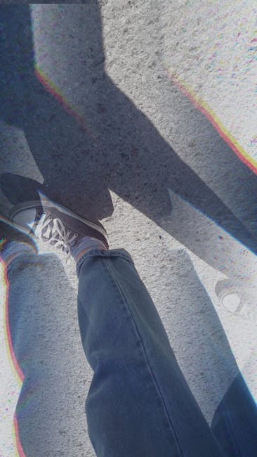 A first-person view from a person standing outside on concrete pavement during a home relocation process, with their left foot visible in a grey sneaker and blue jeans. The person's shadow is cast onto the ground, while nearby, a large cardboard box wrapped in plastic sits on the pavement. In the background, part of a vehicle with an open door and a moving blanket draped over furniture inside the vehicle are visible, indicating a furniture transport and packing process. This scene captures the loading and unloading activities typical of home removals, which might be managed by a professional service like Man with Van Foots Cray, helping clients with efficient packing and furniture transport during a house move on Foots Cray High Street during peak moving day.
