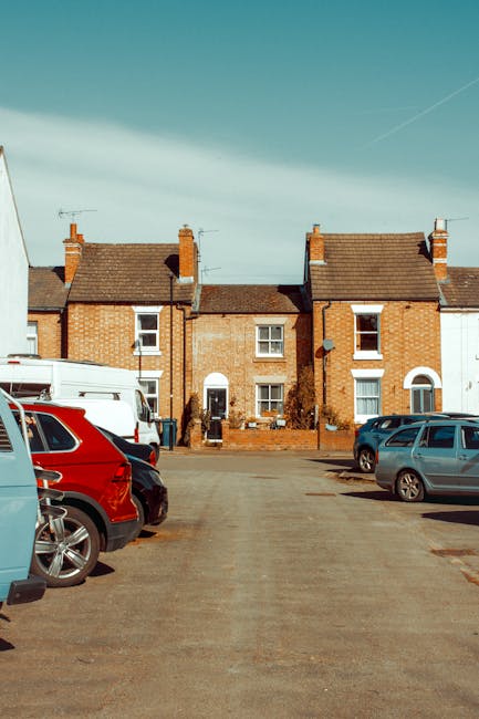 A close-up view of a black and white sneaker worn by a person standing on a paved street surface during a home relocation process. The person is positioned along the white directional arrow painted on the asphalt, indicating a parking or loading zone near a residential property. To the left, there are additional parking lines and shadows cast by nearby objects, possibly vehicles or street furniture. The pavement surface is dark with some small debris and minor cracks. This scene captures part of the loading process for furniture transport, with the arrow guiding the movement of items and vehicles involved in the house removals service provided by Man with Van Foots Cray. The area appears well-lit with natural daylight, and the scene is part of a typical moving day where parking and loading logistics are crucial for efficient home relocation.