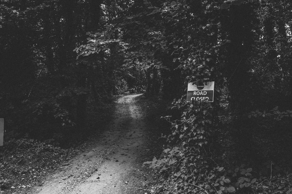 A narrow, unpaved dirt pathway surrounded by dense trees and foliage, with a small sign on the right side that reads 'ROAD CLOSED' attached to a post partially covered by climbing plants. The path app