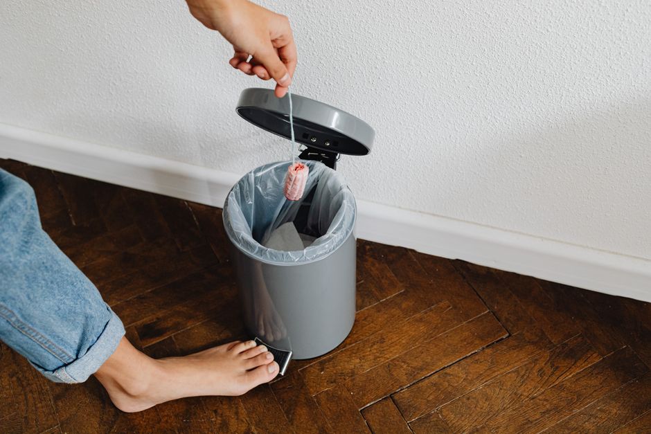 A person's hand tossing a small piece of pink cleaning sponge into a grey plastic waste bin lined with a transparent bag, positioned on a dark wooden floor near a white textured wall. The person is wearing rolled-up blue jeans, with their bare foot visible next to the bin. The scene captures a home environment during packing or decluttering, possibly as part of a house clearance process managed by Man with Van Foots Cray. The waste bin is situated in a corner, and the image reflects typical moving logistics where waste disposal is a part of home relocation or furniture transport steps, aligning with services offered under removals and packing in Foots Cray.