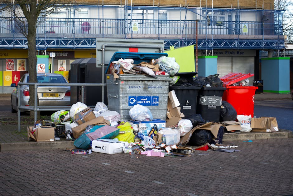 A collection of overflowing waste bins and rubbish bags positioned on a paved street in front of a supermarket with a yellow sign and storefronts in Foots Cray, Bexley. The assortment includes black, red, and grey bins filled with mixed household waste, cardboard boxes, paper, plastic packaging, and ripped cardboard, some of which have spilled onto the ground. The scene is outdoors, with a parked silver car and a metal safety barrier separating the waste area from the pavement, and a building with scaffolding and scaffolding-style fencing visible in the background. The image captures a typical home relocation environment where clearance of household waste is necessary before or after a move, highlighting the importance of adhering to local waste disposal rules applicable in Bexley for Foots Cray jobs, as managed by Man with Van Foots Cray, a professional removals service provider.