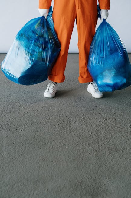 A person's hand tossing a small piece of pink cleaning sponge into a grey plastic waste bin lined with a transparent bag, positioned on a dark wooden floor near a white textured wall. The person is wearing rolled-up blue jeans, with their bare foot visible next to the bin. The scene captures a home environment during packing or decluttering, possibly as part of a house clearance process managed by Man with Van Foots Cray. The waste bin is situated in a corner, and the image reflects typical moving logistics where waste disposal is a part of home relocation or furniture transport steps, aligning with services offered under removals and packing in Foots Cray.