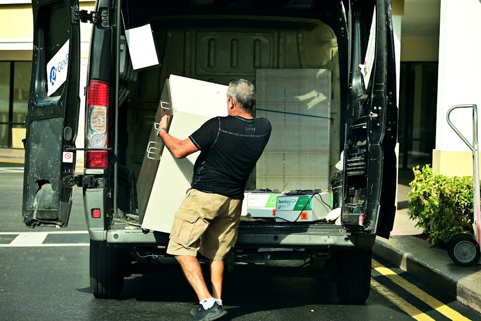 A man with grey hair wearing a black T-shirt, beige cargo shorts, and white sneakers is seen loading a white chest of drawers into the back of a black van parked on a paved area near a building. Inside the van, visible packing materials include cardboard boxes, plastic wrap, and blankets. The unloading process takes place in daylight, with the van opened at the rear doors, and the man is carefully positioning the furniture. To the right, there is a small green shrub and a building with glass windows, indicating an urban or residential setting. The scene illustrates the logistics involved in house removals, furniture transport, and home relocation services offered by Man with Van Foots Cray, highlighting aspects of efficient packing and unloading during a local move near Hall Place.
