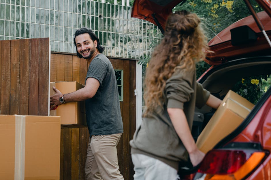 A smiling man with dark hair, wearing a grey t-shirt and beige trousers, is outside a residential property, holding a cardboard box in front of a wooden fence. Next to him, a woman with long curly hair, dressed in a grey jacket, is unloading a cardboard box from the open trunk of a red hatchback car. The vehicle is parked on a paved driveway, and the trunk area contains additional packing boxes. The background features a glass-panelled door and garden greenery. The scene depicts the process of home relocation, with furniture and moving boxes being prepared for transport. The image includes elements such as packing materials, including cardboard boxes, and the use of a car for furniture transport, illustrating the logistics involved in house removals and moving services provided by Man with Van Foots Cray, supporting moving and packing operations near Hall Place.