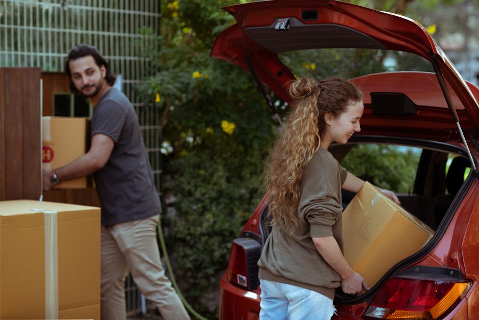 A man with grey hair wearing a black T-shirt, beige cargo shorts, and white sneakers is seen loading a white chest of drawers into the back of a black van parked on a paved area near a building. Inside the van, visible packing materials include cardboard boxes, plastic wrap, and blankets. The unloading process takes place in daylight, with the van opened at the rear doors, and the man is carefully positioning the furniture. To the right, there is a small green shrub and a building with glass windows, indicating an urban or residential setting. The scene illustrates the logistics involved in house removals, furniture transport, and home relocation services offered by Man with Van Foots Cray, highlighting aspects of efficient packing and unloading during a local move near Hall Place.