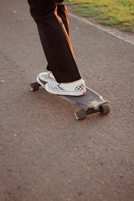 Close-up of a person standing on a skateboard with checkered slip-on shoes, wearing black pants with orange side stripes, on a paved outdoor path. The skateboard features a dark deck with small green decals and black wheels, positioned near a grassy area. This image illustrates casual transport methods commonly used before or during home relocation processes and may relate to the transportation aspect of house removals. Man with Van Foots Cray occasionally incorporates such elements in their furniture transport and packing and moving services, reflecting the movement and logistics involved in local moves, including loading and unloading activities at properties or vehicle loading zones.