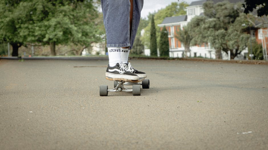 Close-up view of a person's legs wearing white socks with black lettering and black and white Vans sneakers, standing on a skateboard placed on an asphalt street. The background shows a residential area with trees and multi-storey houses, indicating an outdoor urban environment. This scene may relate to activities involved in house removals or the transportation phase of a home relocation, as part of the logistics handled by Man with Van Foots Cray, including transit and outdoor movement preparations.