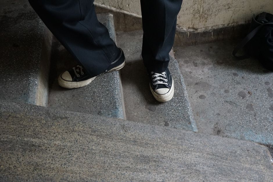 Close-up view of a person wearing black trousers and black-and-white sneakers standing on a staircase with textured concrete steps. The individual appears to be part of a home relocation or furniture transport process. The staircase includes a wooden banister and concrete landing, with packing materials and a black backpack visible nearby. The environment suggests an indoor or semi-indoor space, possibly during the loading or unloading stage of a house move. The focus is on the person's footwear and the stairs, indicative of a moving company's activity, such as Man with Van Foots Cray, performing furniture or item positioning and transportation as part of a professional moving service.