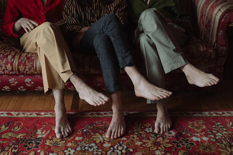 Three individuals sitting on a patterned, floral upholstered sofa inside a residential home, showing only their lower bodies and bare feet resting on a traditional, red-colored rug with intricate designs. The person on the left is wearing beige trousers, the middle person is dressed in dark grey trousers, and the person on the right in light grey trousers. They are seated close together, with their legs slightly apart and feet flat on the wooden floor, in a relaxed position. The sofa has a wooden frame visible beneath the upholstery, and a part of a red curtain or drape can be seen in the background. This scene appears to be set in a living room, with soft lighting, and may be related to a home relocation or packing process, as part of furniture or moving preparations, with no visible moving equipment or boxes in the image. The image is associated with Man with Van Foots Cray, a house removal service specializing in furniture transport and moving logistics.