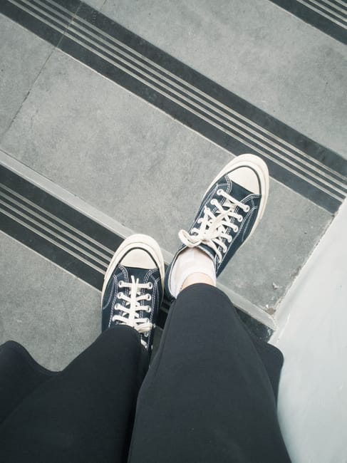 Close-up view of a person wearing black trousers and black-and-white sneakers standing on a staircase with textured concrete steps. The individual appears to be part of a home relocation or furniture transport process. The staircase includes a wooden banister and concrete landing, with packing materials and a black backpack visible nearby. The environment suggests an indoor or semi-indoor space, possibly during the loading or unloading stage of a house move. The focus is on the person's footwear and the stairs, indicative of a moving company's activity, such as Man with Van Foots Cray, performing furniture or item positioning and transportation as part of a professional moving service.