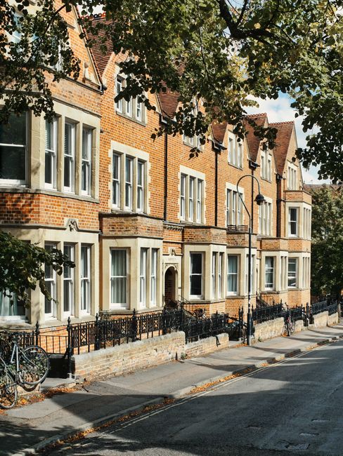 A row of brick residential buildings with multiple white-framed windows and small balconies along a tree-lined street in Foots Cray. The trees provide shade, and fallen leaves are visible on the pavement. A street lamp and black metal fence run parallel to the building facade, separating the sidewalk from the road. In the foreground, a bicycle is parked against the fence. The scene captures a typical urban residential area suitable for house removals and furniture transport, with natural daylight highlighting the architectural details and street environment. This setting aligns with the professional home relocation services offered by Man with Van Foots Cray, emphasizing the importance of careful planning and logistics during packing and moving processes.