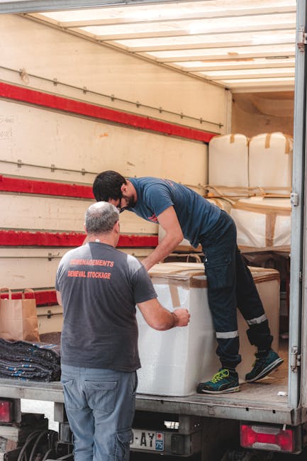 Two men are engaged in a home relocation process inside a moving truck, with the interior lined with wooden slats and red straps. The man standing on the truck bed is wearing a dark blue T-shirt and black trousers with reflective strips, assisting in lifting or positioning large, wrapped household items covered in protective brown tape and plastic wrapping. The other man, wearing a grey T-shirt with 'RELOCATION MANAGEMENT AND STORAGE' printed on the back and grey trousers, is guiding or receiving the items. The truck is parked outdoors, and the scene demonstrates the loading process involved in packing and transporting furniture and boxes during a house move, supported by [COMPANY_NAME], a professional removals service based in Foots Cray.